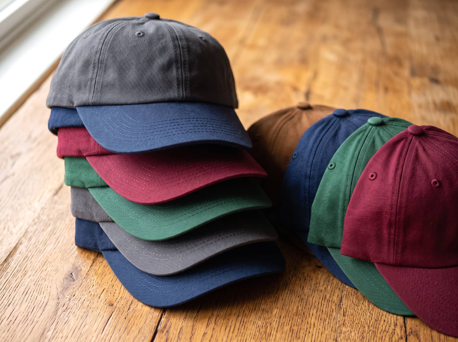 A stack of blank baseball caps in navy, charcoal, green and burgundy.