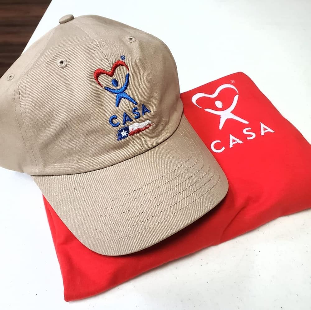 Khaki cap with an embroidered CASA logo beside a red screen-printed shirt carrying the same mark.